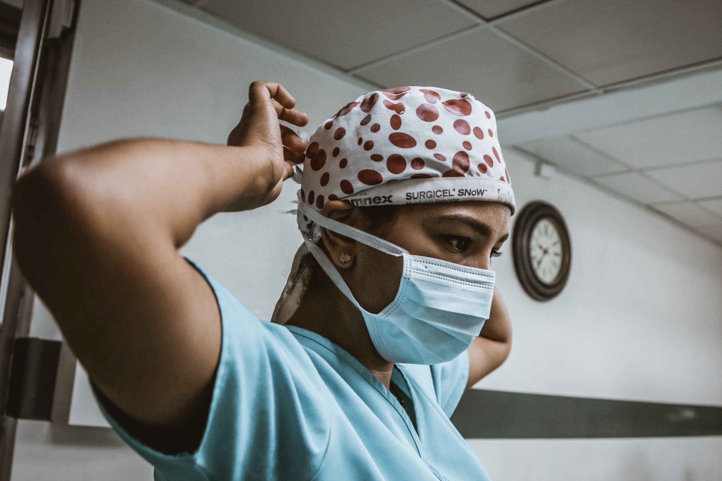 A healthcare professional is adjusting a protective face mask while wearing a polka dot scrub cap, in what appears to be a hospital setting.