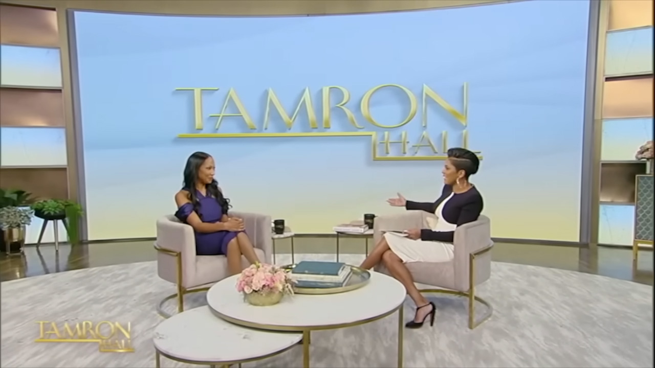Two women are engaged in a conversation on the set of the tamron hall show, seated in a brightly lit studio with stylish decor and the show's logo in the background.