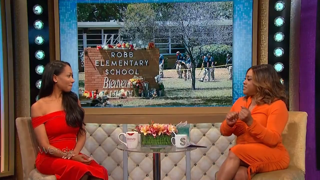 Two women engaged in a conversation on a tv talk show set, with a backdrop screen showing an image of an elementary school and people outside.